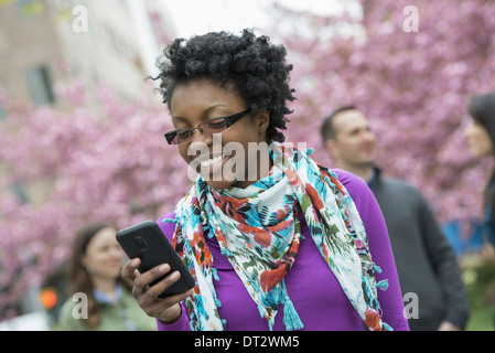 Un gruppo di persone al di sotto dei fiori di ciliegio gli alberi del parco una giovane donna sorridente e controllando il suo telefono Foto Stock