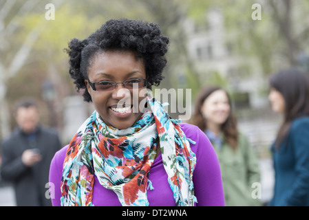 Un gruppo di persone in un parco della città di una giovane donna sorridente indossando una maglietta viola e sciarpa floreale Foto Stock