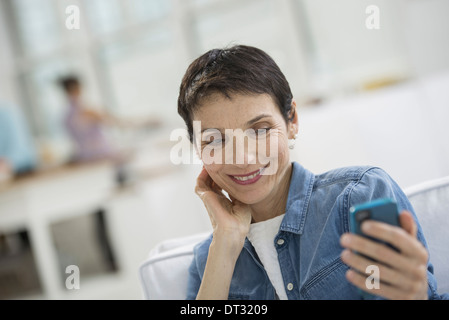 Professionisti in ufficio una luminosa e ariosa e luogo di lavoro di una donna matura in un blu denim shirt guardando un Blu smart phone Foto Stock