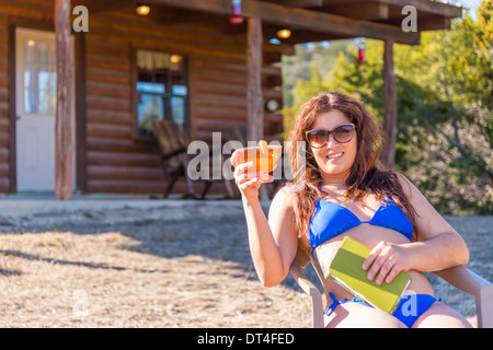 Donna ispanica godersi drink seduto sulla sedia del prato di fronte vacanze home log cabin Foto Stock