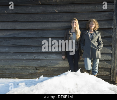 Una madre e figlia in piedi appoggiata contro la parete di legno di un fienile in una fattoria. Neve impilate fino ai loro piedi. Foto Stock