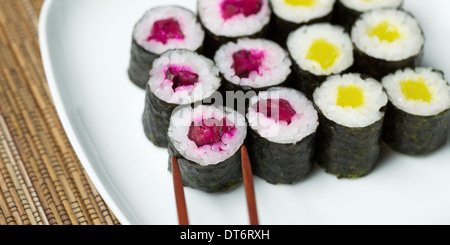 Foto orizzontale di decapare sushi rullo a mano sulla piastra bianca con i bastoncini il prelievo di singoli pezzi e bamboo naturale Foto Stock