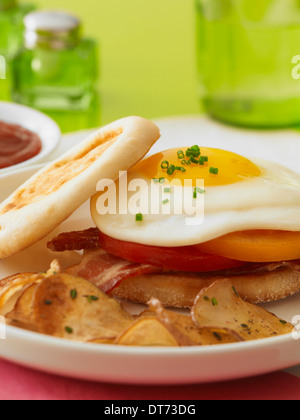 Un'immagine ravvicinata di un soleggiato lato fino a sandwich di uova con pancetta e pomodori in un muffin inglese con prima colazione patate fritte Foto Stock
