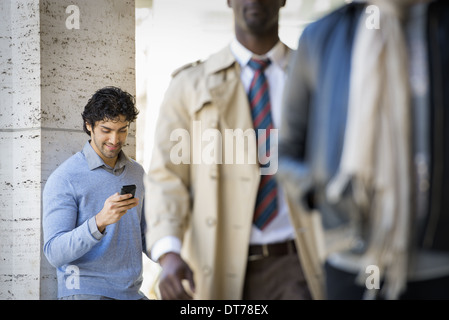 Tre persone sul marciapiede, un uomo con il suo telefono cellulare, un uomo in un impermeabile e un giovane uomo che cammina lungo la strada. Foto Stock