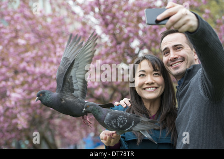 Una coppia di uomo e donna, nel parco, prendendo un selfy, autoritratto con un telefono cellulare. Due piccioni appollaiato sul suo polso. Foto Stock