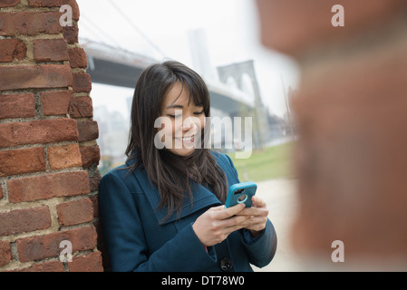 La città di New York. Il Ponte di Brooklyn attraversando la East River. Una donna appoggiata contro un muro di mattoni, controllando il suo telefono. Foto Stock