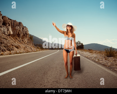 Piuttosto giovane donna con la valigia autostop lungo una strada Foto Stock