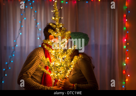 Coppia giovane kissing dietro illuminato albero di natale Foto Stock