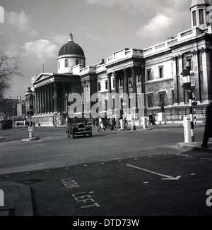 1950, storico, un taxi dell'epoca che viaggia oltre il buidling coperto di fuliggine della National Gallery, Trafalgar Square, Westminster, Londra, Inghilterra, REGNO UNITO. Foto Stock