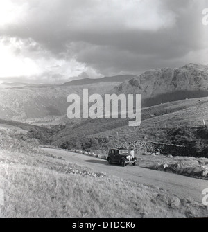 Foto storiche da anni cinquanta che mostra una macchina parcheggiata su una strada di campagna, con il driver all'esterno guardando le opinioni e le colline. Foto Stock