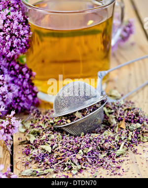 Il tè alle erbe in tazza di vetro, con setaccio in metallo fiori secchi maggiorana, fiori freschi di origano sullo sfondo delle tavole di legno Foto Stock