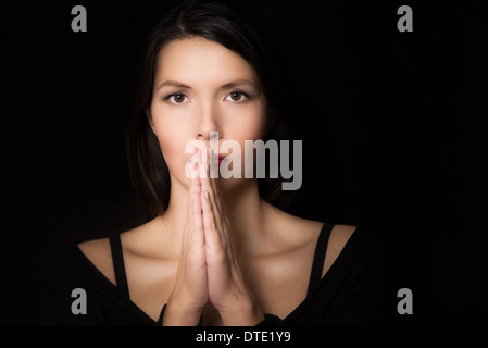Dark suggestivo ritratto di una giovane spirituale donna orante con le mani giunte e un intento guardare negli occhi Foto Stock