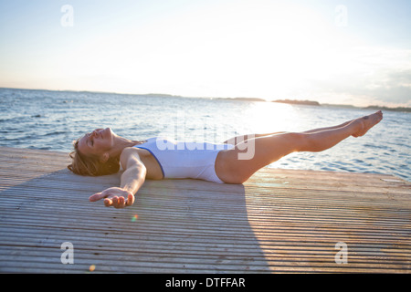 Attraente donna sorridente in bianco costume giacente sul molo in legno nel paese nordico durante l'estate con le gambe sollevate in aria Foto Stock