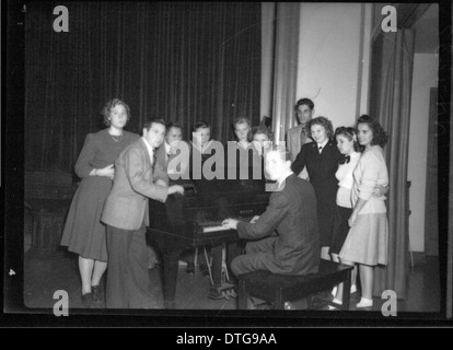 Un ritratto storico di gruppo con personale militare e pianisti riuniti intorno a un pianoforte a coda. Questa fotografia cattura un momento di scambio culturale, mostrando la combinazione di vita militare e performance artistiche in un ambiente sociale. Foto Stock