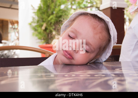 Bambina di due anni di mangiare in caffetterie sulla strada si è addormentato seduto su una sedia e che poggia il capo sul tavolo. Foto Stock