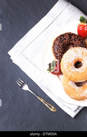 Vista dall'alto sulla piastra di zucchero e ciambelle al cioccolato e fragole fresche servita su grigio tovagliolo tessili su tavola nera. Foto Stock