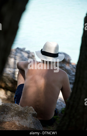 Uomo seduto sulle rocce al mare indossando panama hat visualizzati anche se gli alberi Foto Stock