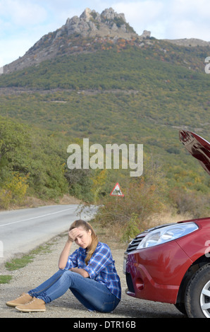 Giovane donna vicino a Broken auto necessita di assistenza seduto sotto il cofano aperto Foto Stock