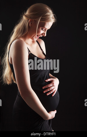 Donna incinta bionda nel terzo trimestre che mostra un'importante scossa del bambino in un abito aderente su uno sfondo nero Foto Stock