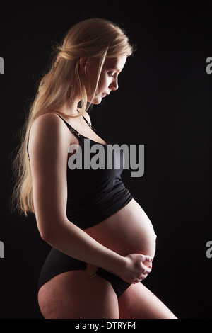Donna incinta bionda nel terzo trimestre che mostra un'importante scossa del bambino in un abito aderente su uno sfondo nero Foto Stock