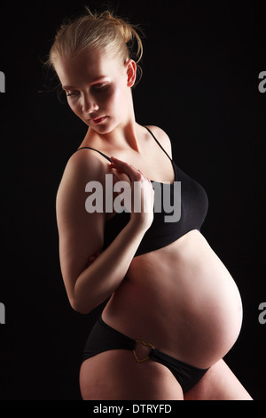 Donna incinta bionda nel terzo trimestre che mostra un'importante scossa del bambino in un abito aderente su uno sfondo nero Foto Stock