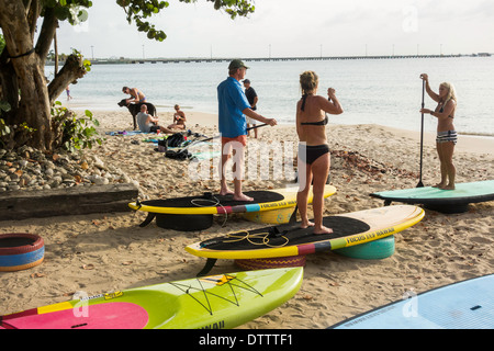 Una donna bionda istruttore insegna una classe in stand up paddle di salire a bordo di un uomo caucasico e la donna nel loro 30s. Sy. Croix, U.S. Isole Vergini. Foto Stock