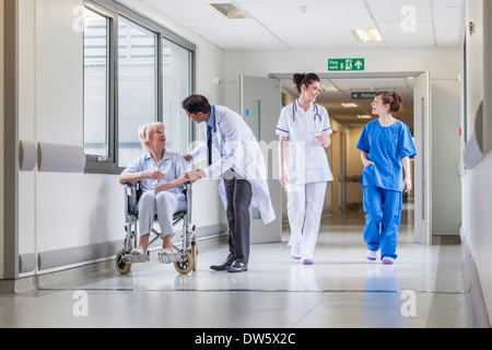 Senior donna femmina paziente in seduta in sedia a rotelle in ospedale corridoio con Asian indiano medico maschio e femmina colleghi infermiere Foto Stock