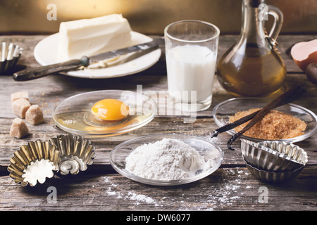 Ingredienti per la cottura (farina, l'uovo, lo zucchero di canna, latte) con l'annata tortina di moduli sul vecchio tavolo in legno. Foto Stock