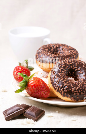 Piastra con ciambelle al cioccolato, fresche fragole e cioccolato fondente servito con tazza di latte sul tessile bianco Foto Stock