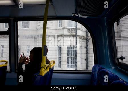 Una solitaria giovane donna si applica il rossetto utilizzando il proprio smartphone come uno specchio durante un bus londinese di viaggio. Foto Stock