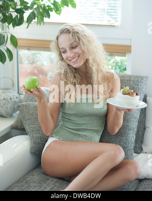 Bella giovane donna di decidere tra torta e Apple mentre è seduto sul divano Foto Stock