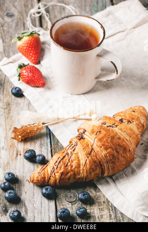 Croissant fresco con la tazza di tè e bacche su un vecchio tavolo di legno. Foto Stock