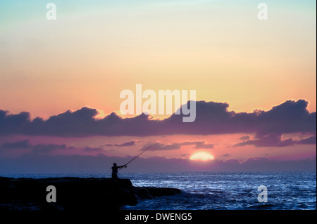 Un solitario rock pescatore getta fuori come il sole sembra da dietro le nuvole all'orizzonte all'alba Foto Stock