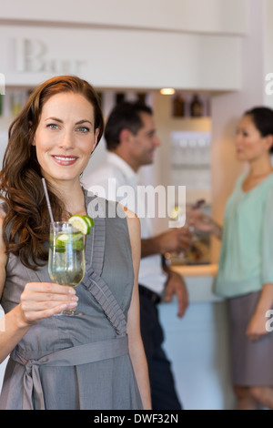 Donna sorridente holding bicchiere da cocktail con gli amici in background in bar Foto Stock
