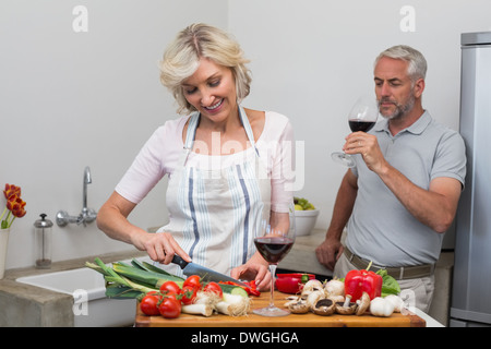 Uomo maturo con bicchiere di vino e la donna tritare verdure in cucina Foto Stock