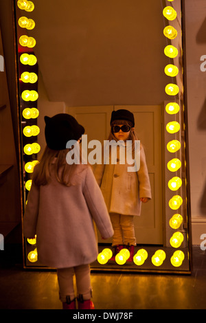 Una ragazzina in occhiali da sole guardando il suo riflesso in uno specchio circondato da lighbulbs Foto Stock