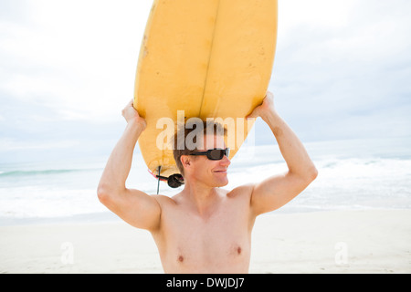 Sorridente surfista maschio in appoggio con la tavola da surf sulla testa alla spiaggia con vista oceano e cielo in background Foto Stock