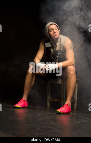 Giovani, boxer aspettando pazientemente per la sua partita seduta tranquillamente su un sgabello di legno in una felpa con cappuccio con le sue mani già bendata Foto Stock