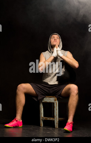 Giovani boxer a pregare con fervore per una vittoria come egli siede in attesa su uno sgabello in legno per l'inizio della sua partita stringendo le mani ed alzare gli occhi al cielo. Foto Stock