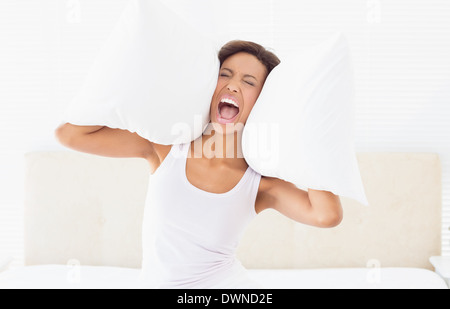 Urlando brunette seduta sul letto che copre le orecchie con cuscino Foto Stock