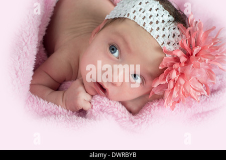Pretty blue eyed baby indossando un archetto di fiori posa su una coperta rosa cercando Foto Stock