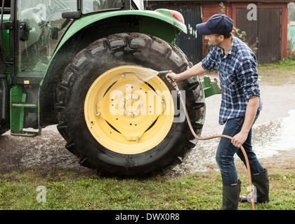 Per tutta la lunghezza della metà adulto agricoltore di lavaggio delle ruote del trattore con il tubo flessibile in una fattoria Foto Stock