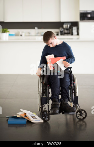 Uomo con paralisi cerebrale guardando caduto libri mentre è seduto su una sedia a rotelle a casa Foto Stock