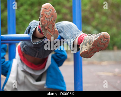 Bambino appeso a testa in giù nel telaio di arrampicata di un parco giochi. Focus sui piedi con solo scarpe. Parti di telaio di arrampicata visibile. Foto Stock