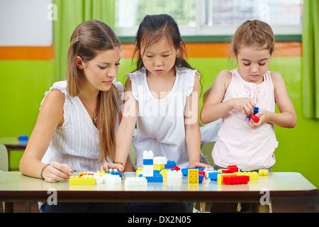 Due bambini che giocano con la costruzione di blocchi in una scuola materna con un insegnante di vivaio aiutando Foto Stock