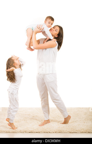 Madre alzando il suo bimbo figlio mentre la figlia di saltare sul tappeto nella loro casa Foto Stock