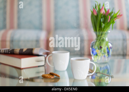 Tazze da caffè sul tavolo di vetro con cucchiaio e biscotti allo zenzero. Prenota e giornale in background Foto Stock