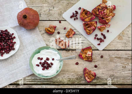 Snack sani una ciotola di yogurt e i semi di melograno. Un frutto intero, semi e pelle su un tagliere, su un tavolo rustico Foto Stock