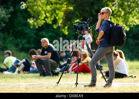 Giovani femmine cameraman in piedi con la sua fotocamera con la folla in appoggio sul prato dietro al festival di musica, ore diurne Foto Stock