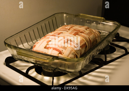 Polpettone crudo avvolto con il bacon in una cesta modello di armatura seduto in un chiaro in Pyrex teglia su una cucina piano cottura Foto Stock